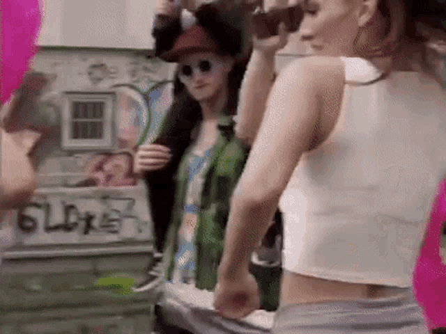 a woman in a white tank top is standing in front of a wall with graffiti on it that says gldx