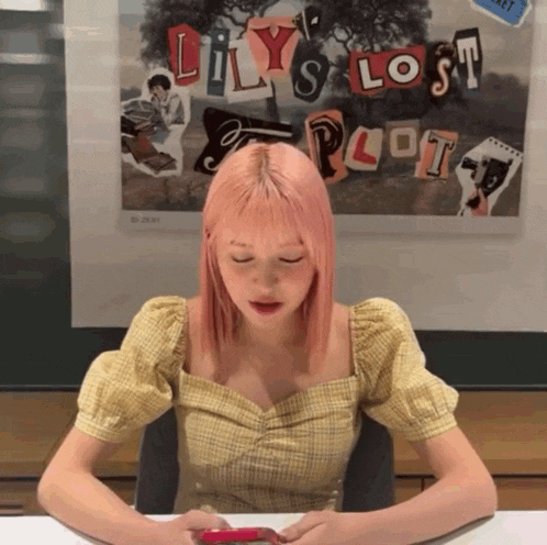 a girl with pink hair is sitting at a desk in front of a poster that says lily 's lost plot