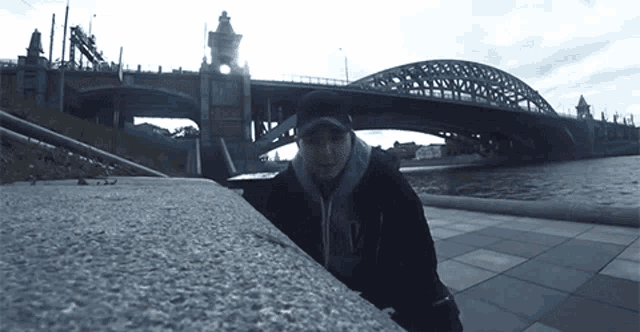 a man in a black hat is standing in front of a bridge over a body of water
