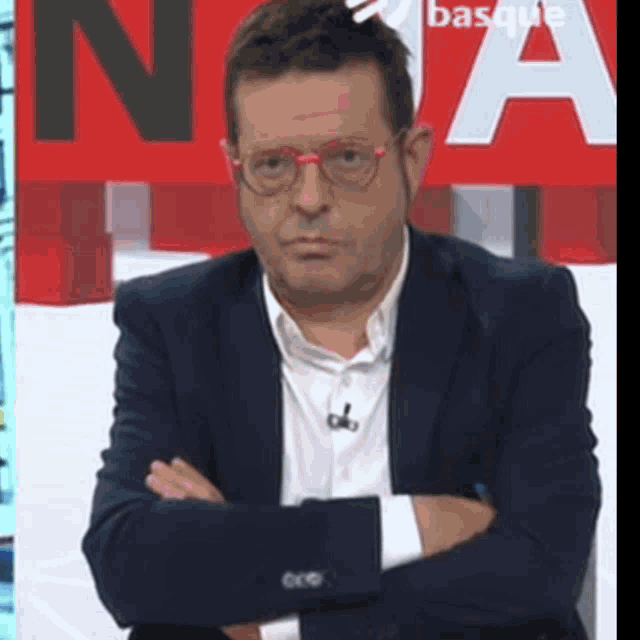 a man in a suit and glasses is sitting with his arms crossed in front of a basque sign