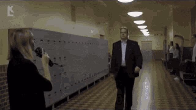 a man in a suit is talking to a woman in a hallway with the letter k on the bottom right