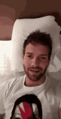 a man with a beard is laying on a bed wearing a white shirt with a picture of a face on it .