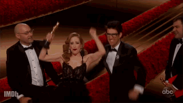 a woman in a black dress is holding an oscar trophy while a group of men in tuxedos cheer