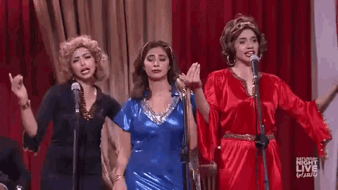 three women singing into microphones on a stage with saturday night live in the background