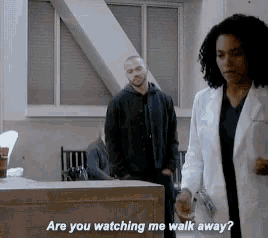 a man and a woman are standing in a room and the woman is asking the man if he is watching her walk away .