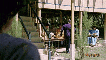 a man is looking at a group of people sitting under a porch with the words wan laksa on the bottom