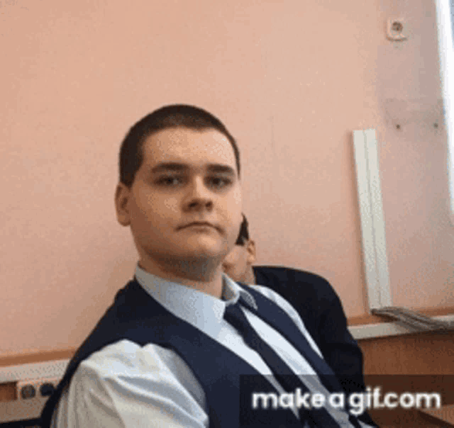 a man in a vest and tie is sitting in front of a pink wall with makeagif.com in the corner