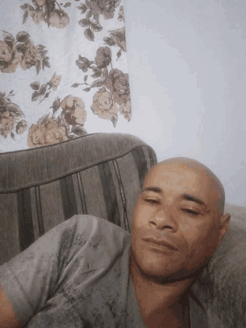 a bald man laying on a couch with a floral curtain in the background