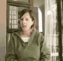 a woman in a green jacket is standing in front of a window and talking .