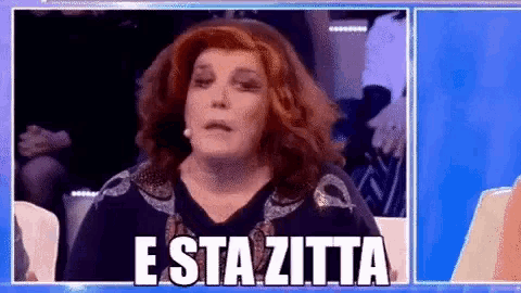 a woman with red hair and a wig is sitting in front of a crowd and says e sta zitta .