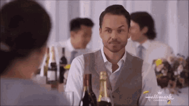 a man in a vest is standing in front of a table with bottles of wine and a woman .