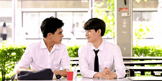 two young men in white shirts and ties are sitting at a table looking at each other .
