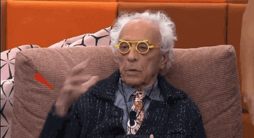 an elderly man wearing yellow glasses and a tie is sitting on a couch .