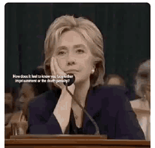 hillary clinton is sitting at a podium with her head resting on her hand .