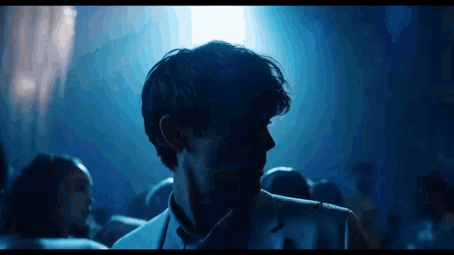a man in a suit and tie is standing in a dark room with a blue light behind him