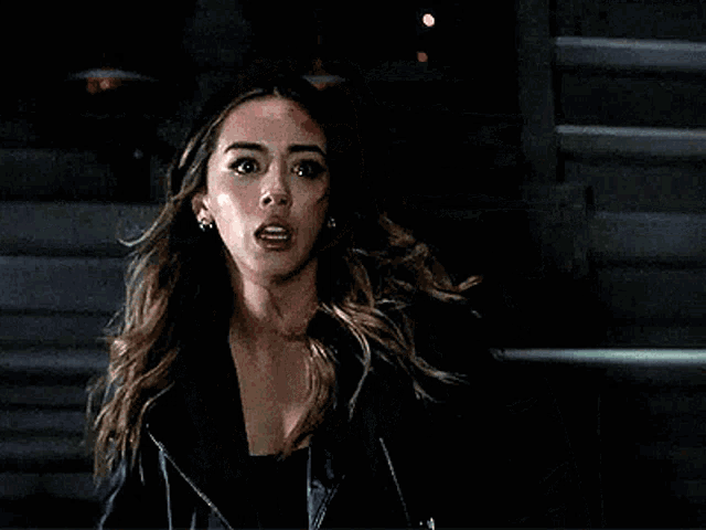 a woman in a black leather jacket with blood on her face is standing in front of a staircase .