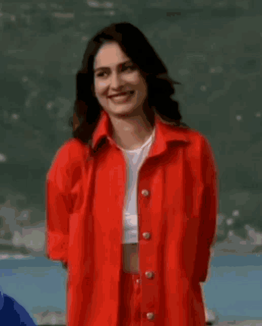 a woman wearing a red jacket and a white crop top is standing in front of a body of water .