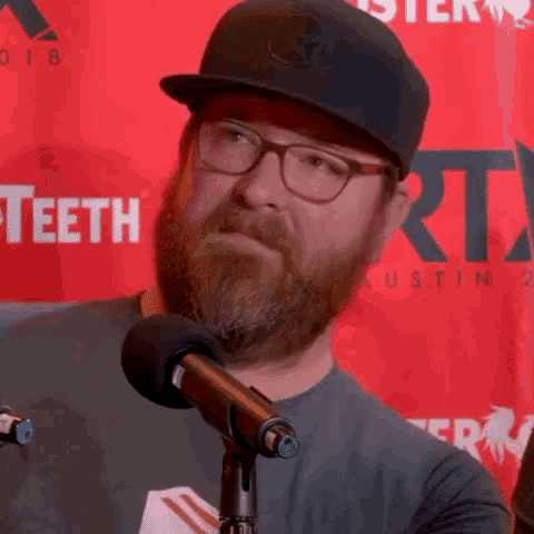 a man with a beard and glasses stands in front of a microphone in front of a red background that says teeth austin