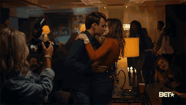 a woman is taking a picture of a man and woman kissing with the bet logo in the background