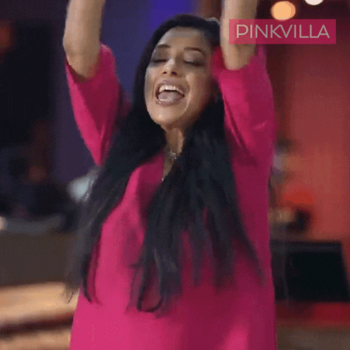 a woman in a pink shirt is raising her arms in the air with a pinkvilla logo behind her