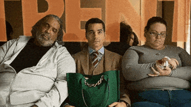 a group of people are sitting in a waiting room . one of the people is holding a green bag .