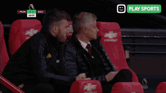 two men are sitting in red chevrolet seats watching a game