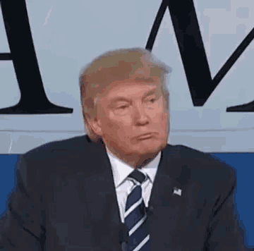 donald trump is wearing a suit and tie and making a funny face while sitting in front of a microphone .