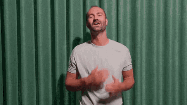 a man in a white t-shirt is standing in front of a green curtain .