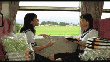 two girls are sitting on a train looking out the window and one is holding a tray of food