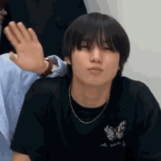 a young man wearing a black shirt and a butterfly necklace is waving his hand .