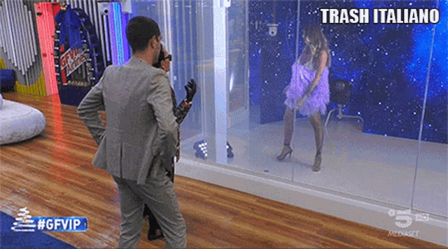 a man in a suit stands next to a woman in a purple dress in front of a glass wall with trash italiano written on it