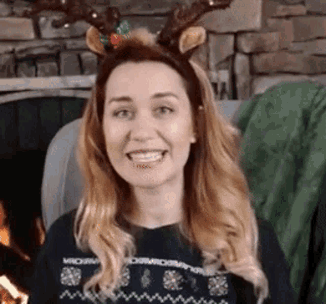 a woman wearing a reindeer headband and a sweater is smiling in front of a fireplace .