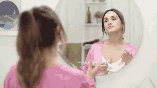 a woman in a pink robe is brushing her teeth in a bathroom mirror