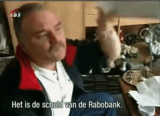 a man with a mustache is sitting at a table with the words het is de schuld van de rabobank