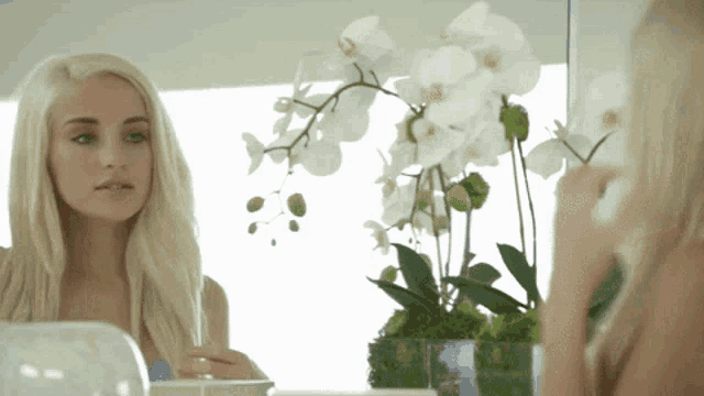 a woman looks at her reflection in a mirror in front of a vase of flowers