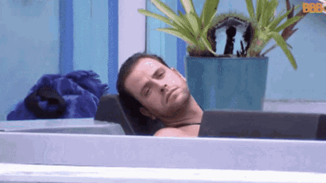 a man is laying in a bathtub with a potted plant in the background and the letters bb on the bottom