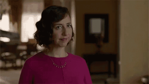 a woman in a pink shirt and necklace is standing in a living room looking at the camera .