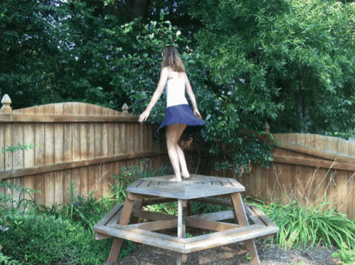 a woman in a blue skirt is standing on a wooden picnic table in front of a wooden fence