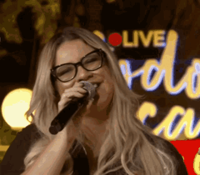 a woman wearing glasses is singing into a microphone in front of a live sign