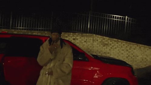a man in a fur coat is standing next to a red car giving the middle finger