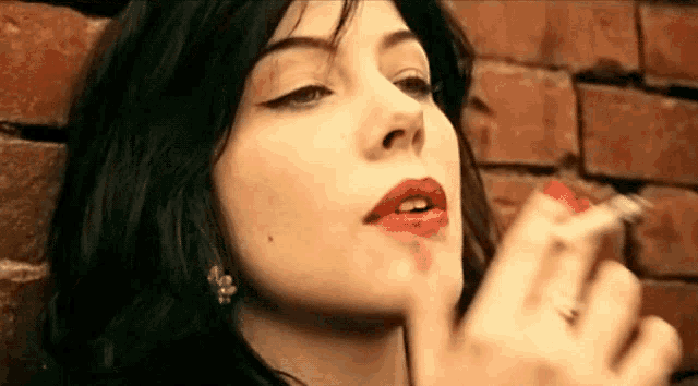 a woman with red lips is smoking a cigarette in front of a brick wall