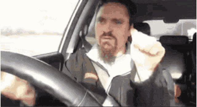 a man with a beard is driving a car and giving the middle finger