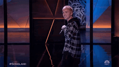 a man in a plaid shirt stands on a stage with a microphone in front of a nbc logo