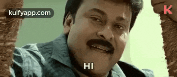 a man with a mustache is standing in front of a brick wall and says hi .