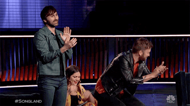 a man in a leather jacket is dancing on a stage while a woman sits on a couch and applauds