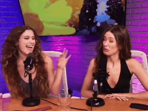 two women are sitting at a table in front of microphones and laughing