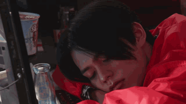 a man in a red jacket is sleeping in front of a cup of ramen
