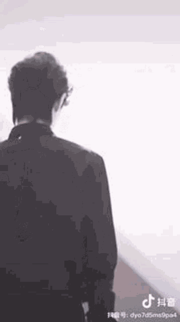 a man in a black suit is standing in front of a white wall and looking at the camera .