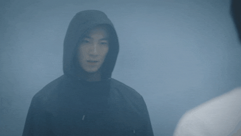 a man wearing a hooded jacket looks at another man in a white shirt