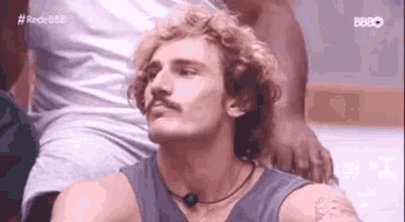 a man with curly hair and a mustache is wearing a gray tank top .